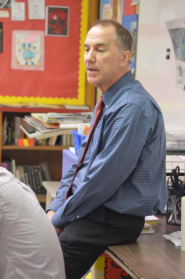 Middle age teacher leaning against a desk in a classroom